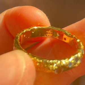 Guldfärgad ring med stenar från Safira - Unik guldfärgad ring från Safira i 925 silver, dekorerad med små färgade stenar i blått, rosa och vitt. Ringen har en cool bubblig textur som ger den en modern och trendig vibe. Perfekt för dig som vill sticka ut med dina accessoarer.