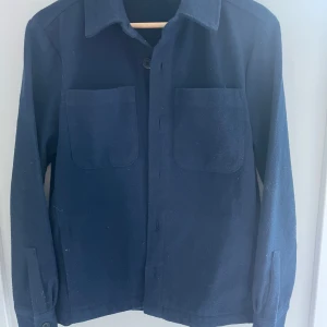 Mörkblå overshirt från Douce De La Sense - Overshirt i mörkblått från Douce De La Sense. Jackan har två stora bröstfickor, klassisk krage och knäppning framtill. Tillverkad i mjukt ullmaterial, perfekt för lager-på-lager. Enkel och clean design som funkar till allt.