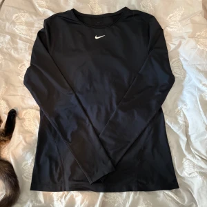 Svart långärmad Nike Dri-Fit tröja - Svart långärmad tröja från Nike med vit logga på bröstet. Tillverkad i Dri-Fit material som andas och transporterar bort fukt, perfekt för träning. Snygg och enkel design med rund hals och normal passform. Storlek M men känns som S