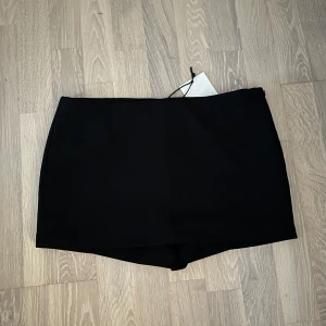 Svart kortkjol med inbygga shorts från Bershka - Enkel och stilren svart kortkjol med inbyggda shorts. Kjolen har en rak passform och är tillverkad i ett mjukt, slätt material. Perfekt att matcha med en crop top eller oversized hoodie för en clean look. Aldrig använd