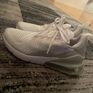 Nike Air Max 270 vita sneakers - Nike Air Max 270 sneakers i vitt med mesh-överdel och stora synliga Air Max-bubblor i hälen. Skorna har snörning, rund tå och detaljer i ljusgrått. Perfekta för dig som gillar sportig och clean stil.