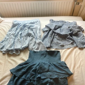 Tre volangkjolar i blå toner - Säljer tre superfina korta kjolar i olika blå nyanser 3 för 50!! . En är enfärgat mörkblå med bred smock i midjan och volanger, en är ljusblå med vitt leopardmönster och volanger, och en är blå med små vita blommor och smock i midjan. Perfekta för sommarens alla tillfällen! 