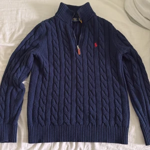 Marinblå kabelstickad tröja Polo Ralph Lauren - Kabelstickad marinblå tröja från Polo Ralph Lauren med halv dragkedja och hög krage. Tröjan har det klassiska röda broderade logot på bröstet och är tillverkad i mjuk ull. Perfekt för dig som gillar en tidlös och preppy stil. Pris 500kr