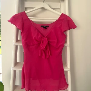 Rosa blus med rosett från H&M - Supersöt rosa blus från H&M i lätt och luftig polyester. Blusen har korta, vingarmliknande ärmar, v-ringning och en stor rosett framtill. Perfekt för dig som vill sticka ut med en färgstark och feminin look.