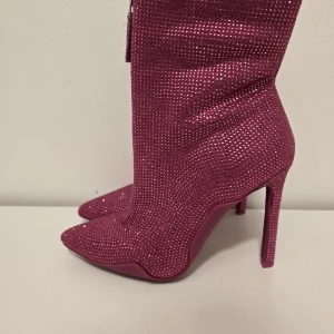 Glittriga rosa stövletter med klack - Snygga stövletter i stark rosa med glittriga stenar över hela skon. De har spetsig tå, hög klack och dragkedja framtill för en edgy look. Perfekta för dig som vill sticka ut och älskar statementskor.