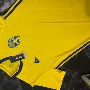 Sverige herrlandslagets matchtröja Adidas - Gul fotbollströja från Adidas med blå detaljer och Sveriges landslagsemblem på bröstet. Klassisk rund hals med blå kant, blå Adidas-logga och blå ränder längs sidorna. Tillverkad i lätt och ventilerande polyester (Climalite). Perfekt för dig som vill visa ditt stöd för Sverige.