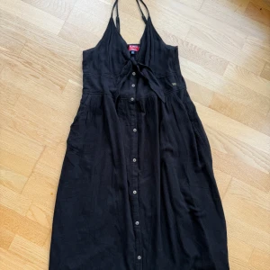 Svart halterneckklänning från Superdry - Svart vadlång klänning från Superdry med halterneck och knyt fram. Klänningen har knappar längs hela framsidan och är tillverkad i ett mjukt, luftigt bomullstyg. Perfekt för varma dagar när du vill känna dig både chill och snygg.