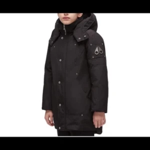Svart parkas Moose Knucklers M junior 12år  - Svart parkas från Moose Knucklers med broderad hockeypatch på insidan och logga på ärmen. Jackan har huva, dragkedja och knappar framtill samt flera fickor. Perfekt för kalla dagar och riktigt snygg streetstil. 