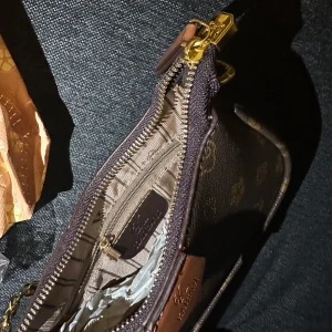Brun axelväska från KAISAQI med kedja - Snygg brun axelväska från KAISAQI med guldiga kedjedetaljer och dragkedja. Väskan har ett monogrammönster i beige och brunt, samt avtagbar axelrem. Tillverkad i syntetmaterial med inslag av skinnimitation. Perfekt storlek för det viktigaste och stilren look.