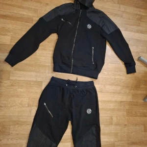 Svarta mjukisbyxor från Philipp Plein - Svarta mjukisbyxor från Philipp Plein med ribbade detaljer på benen och dragkedjefickor. Byxorna har snörning i midjan och en snygg PP-logga framtill. Perfekta för en chill och sportig look.