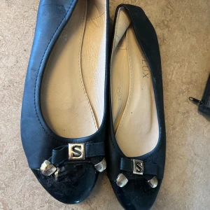 Svarta ballerinaskor med metallrosett - Stilrena svarta ballerinaskor från Lux med rund tå och dekorativ rosett framtill, prydd med metalliska detaljer och ett S i mitten. Skorna har en klassisk design och är tillverkade i skinnliknande material med ljus innersula.