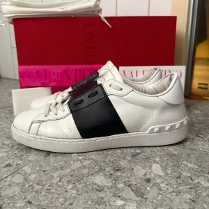 Valentino Open - Stilrena Valentino Garavani Open sneakers i vitt skinn med bred svart läderdetalj över mitten. Klassisk rund tå, vita snören och ikoniska nitar på hälen. Grymt skick! de är väldigt vita, med väldigt lite slitningar och nästan bara smutsiga under vilket varken märks eller syns under användning. Självklart äkta! Köpta i butik och alla tillbehör såsom box, två st dustbags, fyra st valentino kort och även kvitto finns och medföljer vid köp! Storlek 42, passar 42/43. Frågor? Bara att höra av sig!