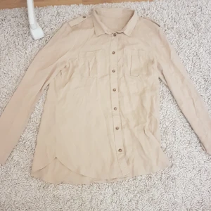 Beige skjorta med fickor och axelremmar - Stilren beige skjorta med långa ärmar, två bröstfickor och snygga axelremmar med knappdetalj. Skjortan har klassisk krage och knäppning framtill. Perfekt för en clean och avslappnad look. PRIS KAN DISKUTERAS!