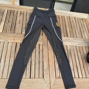 Svarta Maya Delorez ridleggings - Svarta ridleggings från Maya Delorez med hög midja, gråa sömmar och silikonprint längs insidan av benen för extra grepp. Stretchigt material och praktiska fickor på sidorna. Gult MD-logga på låret. Perfekta för ridning och träning.