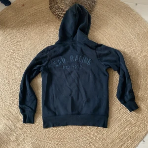Svart hoodie från Sail Racing - Svart hoodie från Sail Racing med broderad logga och text på ryggen. Tröjan har dragkedja framtill, huva och långa ärmar med muddar. Perfekt för dig som gillar en stilren och sportig look. Materialet känns mjukt och bekvämt, troligen bomullsmix.