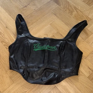 Svart korsett-topp från SHEIN - Cool svart korsett-topp i skinnimitation från SHEIN med grönt broderi 'Visual feast' framtill. Toppen har breda axelband, figurnära passform och dragkedja i ryggen. Perfekt för att lyfta vilken outfit som helst med edgy vibe.