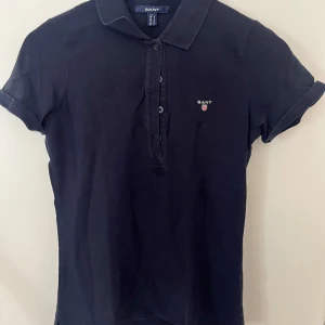 Svart pikéblus från Gant - Klassisk svart pikéblus från Gant med korta ärmar, krage och knappar framtill. Liten broderad Gant-logga på bröstet. Perfekt för en clean och sportig look. Gjord i mjuk bomull som andas. 1 kostar 180kr