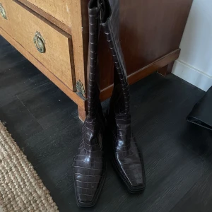 Bruna stövlar med krokodilmönster - Snygga höga stövlar i mörkbrunt skinn med krokodilpräglat mönster och fyrkantig tå. Stövlarna har en klassisk siluett och dragkedja på insidan. Perfekta för att lyfta din outfit med en cool och trendig touch.