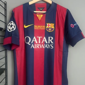 FC Barcelona Messi 10 Champions League tröja - Säljer en officiell FC Barcelona fotbollströja från Champions League-finalen 2015 med Messi 10 på ryggen. Tröjan är randig i mörkblått och rött, har korta ärmar och är tillverkad i lätt, ventilerande Dri-Fit-material. Tryck med Qatar Airways, klubbmärke och Champions League-logga. Helt ny med tagg en replika