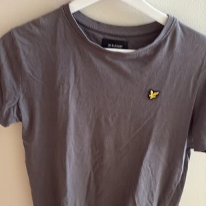 Grå t-shirt från Lyle & Scott - Snygg grå t-shirt från Lyle & Scott med klassisk rund hals och korta ärmar. På bröstet sitter den ikoniska gula örnloggan som ger en clean och stilren look. Tillverkad i mjuk bomull som är skön att ha på sig hela dagen.