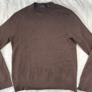 Brun stickad tröja Polo Ralph Lauren - Brun stickad tröja från Polo Ralph Lauren i ylle men känns som Kashmir för den är väldigt mjuk o skön, med rund hals och grönt broderat logotyp på bröstet. Klassisk modell med ribbade muddar och lång ärm, perfekt för lager-på-lager under kyliga dagar. Den har ett litet hål som inte syns vid användning men annars toppskick👌