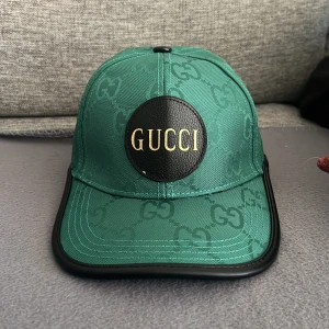 Gucci keps - Snygg grön keps från Gucci med GG-monogram över hela och stor svart patch med guldig GUCCI-text framtill. Kepsen har justerbar rem bak och detaljer i svart skinn. Tillverkad i polyester och skinn, perfekt för dig som vill sticka ut.