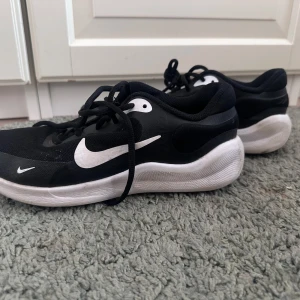 Svarta Nike sneakers strl 37,5 - Svarta Nike sneakers med vit sula och stora vita swoosh-loggor på sidorna. Ovandelen är i mesh och syntet, vilket gör dem lätta och luftiga. Klassisk snörning och rund tå. Perfekta för dig som gillar sportig stil och vill ha bekväma skor till vardagen.