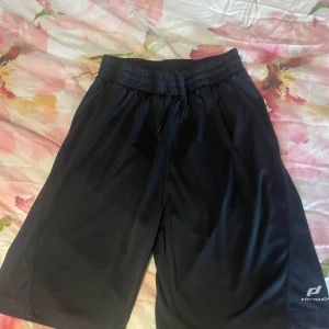 Svarta träningsshorts Pro Touch - Svarta träningsshorts från Pro Touch med elastisk midja och snörning. Lätta och luftiga i syntetmaterial, perfekta för sport eller gym. Klassisk design med logga på benet och normal passform.