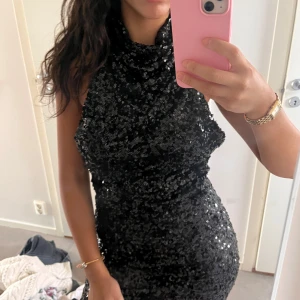 Svart glittrig paljettklänning - Supersnygg svart kort paljettklänning med hög hals och ärmlös design. Klänningen har en tight passform och är täckt av glittrande paljetter som verkligen fångar ljuset. Perfekt för dig som vill sticka ut och glänsa på nästa fest!