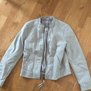 Ljusgrå skinnjacka från Vero Moda - Snygg ljusgrå skinnjacka från Vero Moda med dragkedja framtill och två fickor. Jackan har en stilren design med markerade sömmar och lätt krage. 