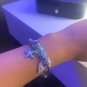 Silverfärgat armband med berlocker - Snyggt silverfärgat armband med flera berlocker, bland annat en blå sjöstjärna, glittrigt hjärta och blommor i blått och silver. Armbandet har även en turkos pärla och detaljer som ger en fräsch och somrig vibe. Perfekt accessoar för att lyfta din outfit.