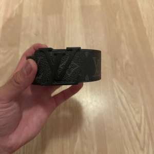 Svart Bälte - Säljer ett svart L bälte med ikoniskt L spänne i matt svart metall. Bältet är brett och har diskret monogrammönster på lädret. Insidan är brun och materialet känns lyxigt och robust. Perfekt för dig som vill ha en statement-accessoar.