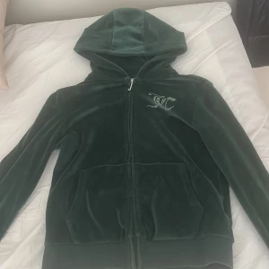 Juicy Couture grön velour hoodie - Mörkgrön velour hoodie från Juicy Couture med glittrande strasstenar på bröstet och stor Juicy Couture-logga på ryggen. Jackan har dragkedja, huva och två fickor framtill. Perfekt för dig som vill ha den ikoniska Y2K-looken.