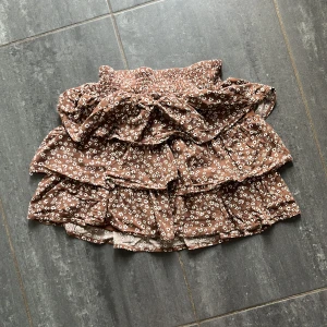 Brun blommig volangkjol från Lindex - Supersöt brun kortkjol från Lindex med vita småblommor och flera volanger. Kjolen har bred resår i midjan för skön passform och är perfekt för dig som gillar en lekfull och trendig stil. 