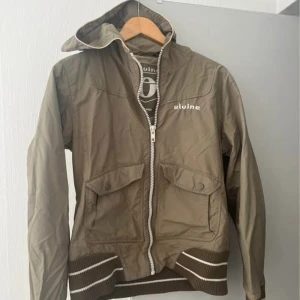 Olivgrön vindjacka från Elvine L - Olivgrön vindjacka från Elvine med huva, dragkedja och två stora fickor framtill. Jackan har ribbade muddar med vita ränder och en sportig vibe. Perfekt för blåsiga dagar och enkel att matcha med jeans eller sneakers.