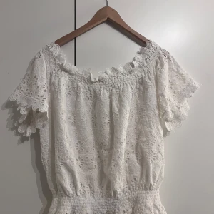 Vit spetsblus från Lindex x By Malina - Superfin vit blus med broderade blommor och spetsdetaljer från Lindex x By Malina. Blusen har offshoulder-modell, korta ärmar med volang och smock i midjan. Perfekt för sommarens alla tillfällen!