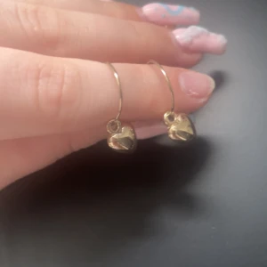 Guldiga örhängen med hjärtanhängen - Snygga örhängen i tunn guldfärgad metall med små blanka hjärtanhängen. Perfekta för dig som gillar söta detaljer och vill ha något som sticker ut men ändå är stilrent. Passar till alla tillfällen och ger en gullig touch till din look.