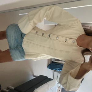 Beige jeanstopp med vida ärmar - Snygg beige jeansjacka med markerade sömmar och vida ärmar. Jackan har rund halsringning och coola silverfärgade knappar framtill. 