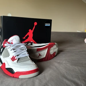 Nike Air Jordan 4 Fire Red sneakers - Nike Air Jordan 4 Fire Red sneakers med vit ovandel i skinn, röda och svarta detaljer samt meshpaneler på sidorna. Klassisk Jumpman-logga på plösen och hälen. Snygg design med synlig Air-enhet i sulan och coola plastdetaljer vid snörningen.