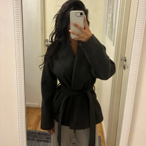 Mörkgrå omlottjacka från ONLY - Snygg mörkgrå omlottjacka från ONLY med bred krage och bälte i midjan. Jackan har en stilren och enkel design, perfekt för dig som gillar en clean look. Tillverkad i ullblandning och har långa ärmar. Passformen är normal och jackan är helfärgad utan mönster.