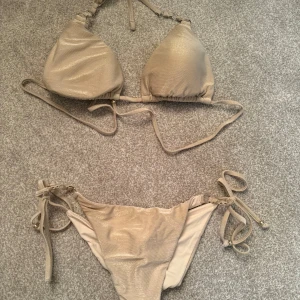 Champagnefärgad bikini med knyt - Snygg champagnefärgad bikini med glansig finish. Triangeltopp med knyt i nacken och ryggen, samt matchande bikinitrosa med knyt i sidorna och rynkade detaljer bak. Perfekt för stranden eller poolen, ger en lyxig vibe. Storlek S