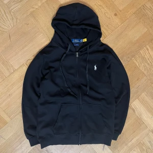 Svart hoodie från Polo Ralph Lauren - Svart hoodie från Polo Ralph Lauren med vit broderad logga på bröstet. Tröjan har dragkedja, huva med snörning och två fickor framtill. Tillverkad i en mjuk blandning av bomull och polyester. Perfekt för en chill och stilren look.