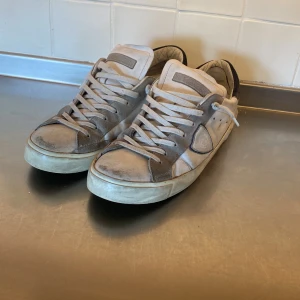 Vita och grå sneakers från Philippe Model - Snygga sneakers från Philippe Model i vitt och grått med klassisk låg profil. Skorna har snörning, rund tå och detaljer i mocka och skinn. Märkets logga syns på sidan. Perfekta för en avslappnad och trendig look.