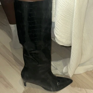 Svarta stövlar med krokopräglat mönster - Snygga svarta stövlar i skinnimitation med krokopräglat mönster och spetsig tå. De har låg klack och en glansig finish som ger en cool och trendig vibe. Perfekta att styla med kjol eller jeans för en edgy look.