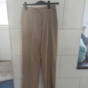 Beiga kostymbyxor med raka ben - Snygga beiga kostymbyxor från Nakd med raka ben och klassisk passform. Byxorna har hällor för bälte, diskreta sidofickor och är tillverkade i ett mjukt material som känns skönt mot huden. Perfekta för en clean och stilren look. Köparen står för frakten. 