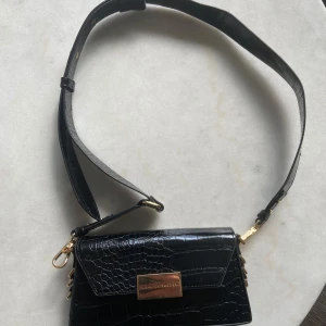 Svart axelväska från iDeal of Sweden - Snygg svart axelväska från iDeal of Sweden med croco-mönstrad yta och guldfärgade metalldetaljer. Väskan har ett brett justerbart axelband och ett elegant lås framtill. Perfekt storlek för det viktigaste och med innerfack för extra organisering.