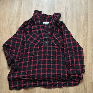 Rutig henley-skjorta från Isolde - Svart och röd rutig henley-skjorta från Isolde med trekvartsärm och knappar framtill. Skjortan har en avslappnad passform och är tillverkad i mjuk bomull. Sparsamt använd + inga synliga defekter (utom något nopprig)!!!