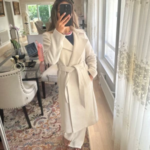 Beige kappa med bälte och slag - Snygg beige kappa med klassiska slag och bälte i midjan. Lång modell med knappar framtill och slits bak. Perfekt för dig som vill ha en stilren och tidlös look. Passar till allt och är superenkel att styla. Använd men ej trasig eller sliten. Tjocktröja får plats under