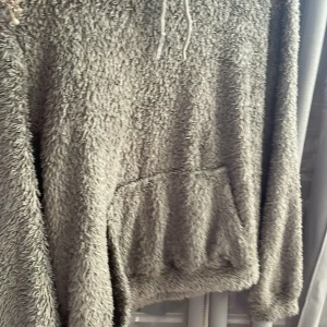 Mysig grå fluffig hoodie - Supermjuk och fluffig grå hoodie med huva och dragsko. Tröjan har en stor magficka och långa ärmar, perfekt för kalla dagar. Materialet är teddy/fleece-liknande och ger en riktigt cozy vibe.