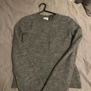Grå stickad tröja  - Mysig grå stickad tröja med rund hals och långa ärmar. Tröjan har ribbade muddar vid ärmslut och nederkant, vilket ger en snygg och enkel look. Perfekt att slänga på sig när det är kyligt ute.